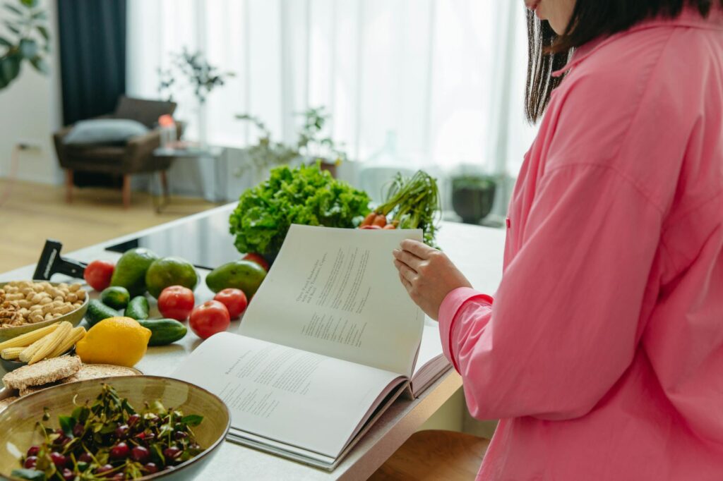 A woman in a kitchen flipping through a cookbook surrounded by fresh vegetables.